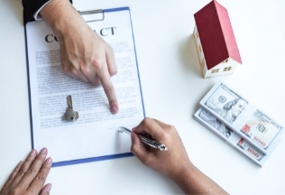 Hands signing a property contract with house model, keys, and cash — NRIWAY NRI property and legal documentation services.